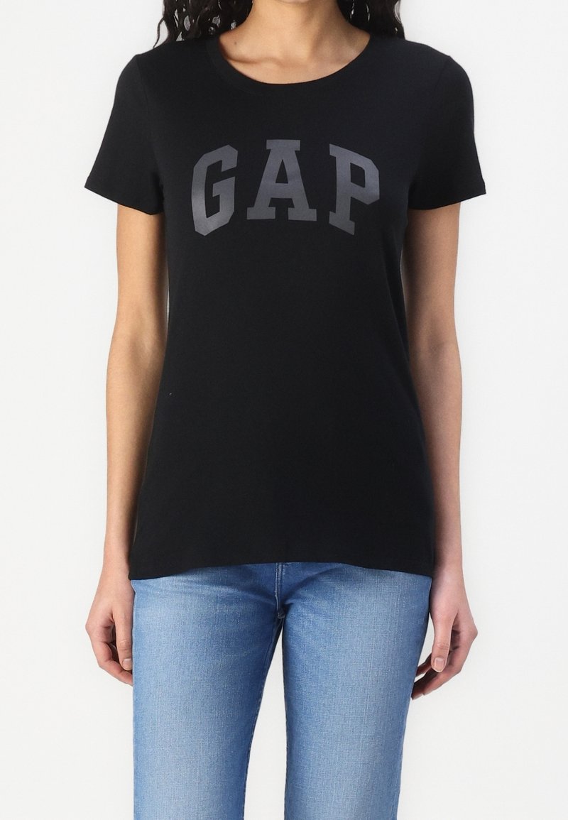 Femme portant un t-shirt noir à manches courtes avec logo GAP gris foncé et un jean bleu clair sur fond blanc.