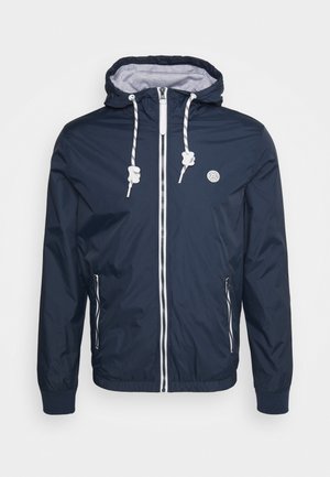 Veste coupe-vent marine avec capuche, poignets élastiques et fermeture éclair sur le devant. Comprend des cordons de serrage et une doublure intérieure grise avec un détail de logo.