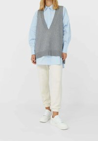 Personne portant une chemise boutonnée bleu clair, un gilet sans manches en tricot gris, un pantalon de jogging crème et des baskets blanches, se tenant sur un fond blanc.
