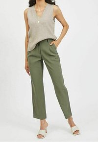 Světlobéžový top bez rukávů s véčkovým výstřihem, spárovaný s olivově zelenými elegantními kalhotami. Modelka nosí světlé sandály a vrstvené zlaté náhrdelníky.