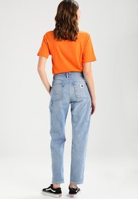 Orange kortärmad t-shirt tillsammans med ljusblå högmidjade jeans med två bakfickor och en logotyppatch. Svarta sneakers synliga.