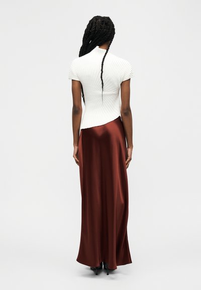 Femme aux longs cheveux tressés portant un haut blanc à manches courtes côtelé et une longue jupe en satin marron brillant, debout de dos.