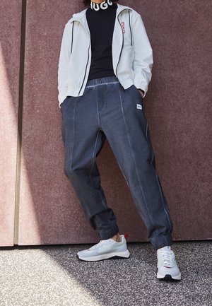 Lichtgrijs jack met rits en capuchon met trekkoord, zwarte coltrui, ruimvallende donkergrijze broek en lichtgekleurde sneakers met rode accenten.