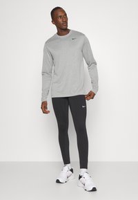 Γκρι μακρυμάνικο μπλουζάκι με μαύρο λογότυπο, σε συνδυασμό με μαύρα κολάν και λευκά αθλητικά παπούτσια με μαύρες και κόκκινες λεπτομέρειες. Ομαλή υφή.