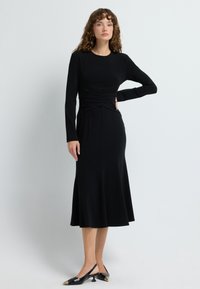 Schwarzes langärmliges Kleid mit einer taillierten Oberkörperpartie und Raffungsdetails sowie einem ausgestellten Rock. Kombiniert mit schwarzen Slingback-Schuhen mit goldenen Akzenten.