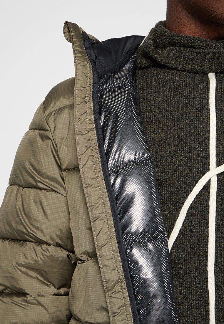 Chaqueta acolchada verde con un forro negro brillante. El exterior tiene un acabado texturizado y un patrón de acolchado visible. Debajo lleva un suéter de punto oscuro.