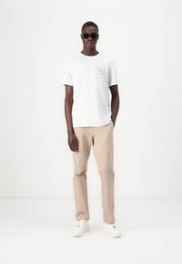 Hvit bomull t-skjorte med "GUESS" logo på brystet, beige bukser og hvite joggesko. Modell iført svarte solbriller, står mot en lys bakgrunn.