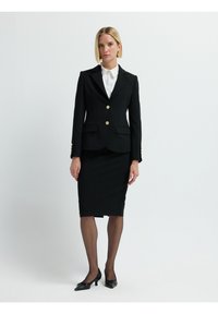 Blazer negro con botones dorados, corte entallado, combinado con una falda negra hasta la rodilla. Una camisa blanca de cuello y pantys negros transparentes completan el conjunto.