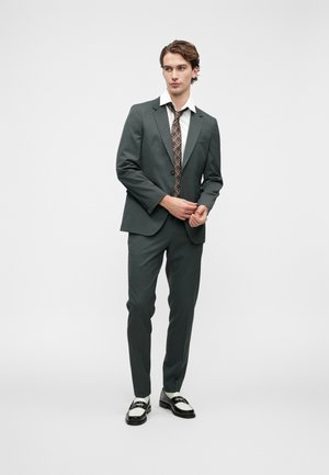 Mørkegrøn dragt med en tætsiddende blazer, matchende bukser og en hvid skjorte med et mønstret slips. Sorte og hvide sko fuldender outfittet.