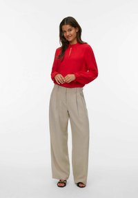 Mujer de pie vistiendo una blusa roja de manga larga, pantalones de pierna ancha beige y tacones negros abiertos por delante, contra un fondo blanco.