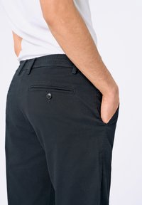 Pantalons noirs avec une texture lisse, une taille ajustée, deux poches arrière avec fermetures à bouton et un design épuré.