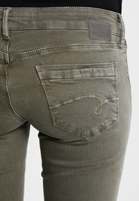 Olivově zelené džíny z denimu s mírným vzestupem, které mají koženou cedulku s logem na zadní straně a výrazné prošívání na kapsách.