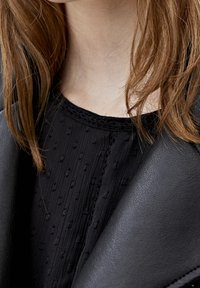 Cou du torse et poitrine d'une femme avec des cheveux brun rougeâtre, portant un chemisier noir texturé et une veste en cuir noire.