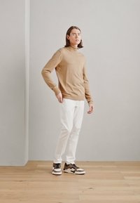 Maglione a collo alto di colore marrone chiaro, pantaloni bianchi e scarpe da ginnastica con accenti neri e marroni. Pavimento in legno e sfondo grigio uniforme.