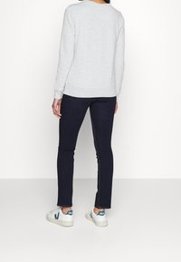 Personne portant un sweat-shirt gris clair, un jean slim bleu foncé et des baskets blanches VEJA, debout de dos face à la caméra.