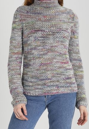 Hrubý pletený rolák ze šedé směsové příze s barevnými skvrnkami, nošený s modrými džínami, zobrazený od krku po boky.