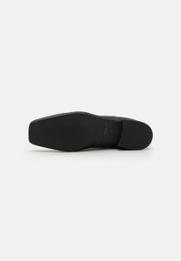 Suela de zapato de cuero negro, diseño plano con tracción de goma texturizada, punta redondeada y bordes limpios. Presenta el nombre de la marca grabado en la suela.