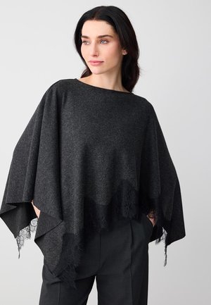 Frau trägt einen dunkelgrauen Poncho mit Spitzenbesatz und schwarze Hose, schaut leicht zur Seite und hat die Hände in den Taschen.