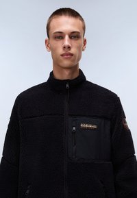 Schwarze Fleecejacke mit einem Reißverschluss auf der Vorderseite, strukturiertem Äußeren und einer kontrastierenden Tasche mit einem Logopatch. Mit seitlicher Reißverschlusstasche.