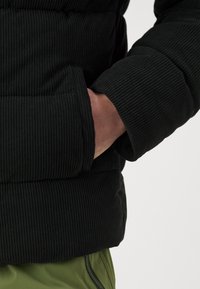 Černá kordurová bunda s ribbovanou texturou, která má kapsu a manžetový rukáv. Bunda je nošena přes olivově zelené kalhoty.