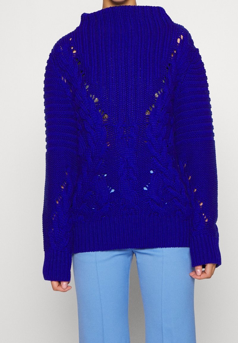Personne portant un pull en grosse maille bleu vif avec des motifs torsadés et un pantalon bleu clair, debout devant un fond blanc uni.