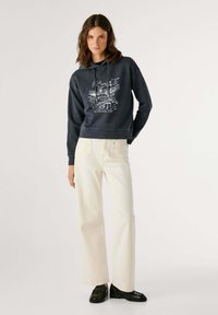 Felpa con cappuccio blu navy con stampa grafica bianca sul davanti, abbinata a jeans a gamba larga beige chiaro e scarpe nere, caratterizzata da una texture liscia.