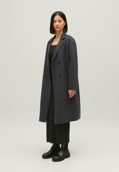 Manteau en laine gris foncé croisé avec de grands revers, coupe décontractée et poches latérales, associé à une robe en maille noire et des bottines noires.