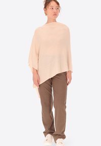 Poncho lavorato a maglia beige con una vestibilità ampia, caratterizzato da una consistenza morbida. Abbinato a pantaloni a righe marroni e sneakers bianche.