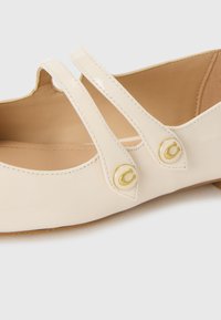 Beige patent leather shoe with two narrow straps, gold circular buttons featuring a "C" accent, and a rounded toe design.