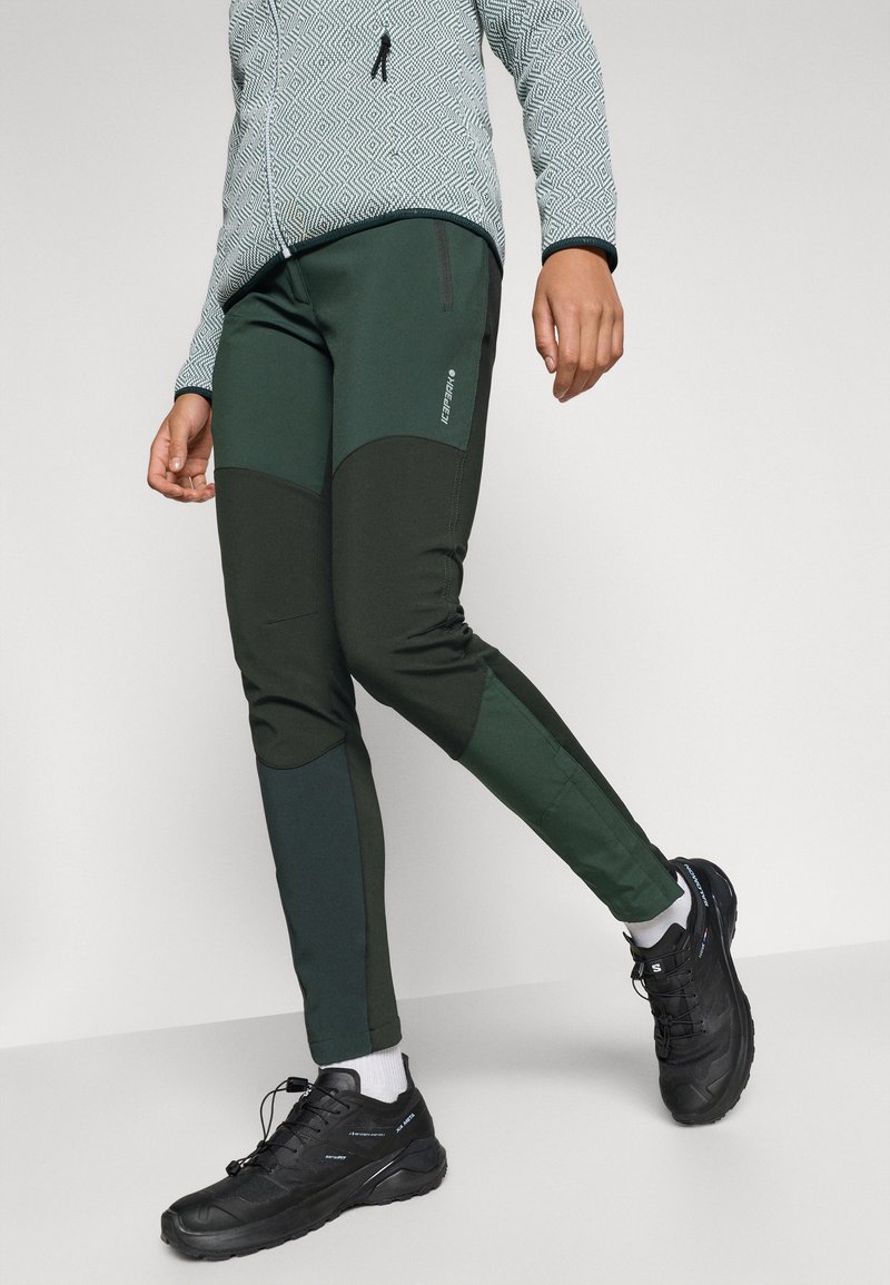 Zielone spodnie sportowe z kontrastującymi ciemnymi panelami, zwężanym krojem i gładką fakturą, w połączeniu z czarnymi sportowymi butami i białymi skarpetkami.