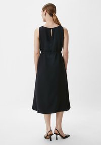 Schwarzes ärmelloses Kleid mit einem Schlüssellochdetail am Rücken, fließendem Stoff und Knöchellänge. Kombiniert mit schwarzen High Heels.