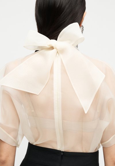 Femme portant un chemisier crème transparent avec un grand nœud noué à l'arrière du cou et un bas noir taille haute, vue de dos.