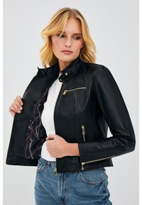 Black leather jacket with a high collar, zippered pockets, and gold hardware. Interior features a patterned lining. Worn over a white top.