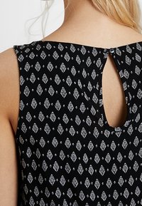 Blusa nera con un motivo geometrico bianco. Presenta un colletto rotondo e un piccolo apertura a fessura sul retro, chiusa con un bottone.
