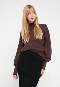 Brązowy sweter z dzianiny z wysokim kołnierzem i luźnymi rękawami, zestawiony z czarną ołówkową spódnicą prążkowaną, z delikatnym zaokrąglonym wykończeniem na dole.