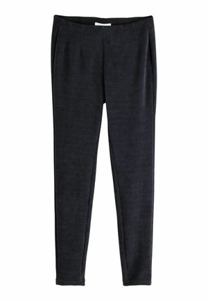 Kvinde iført mørkstribet top, mørke leggings, røde flade sko og solbriller står på trægulv med beige stofbaggrund.