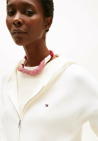 Witte rits-hoodie met een klein logo op de linkerborst, gedragen over een textuur crème top, aangekleed met een rood en witte gedraaide halsdoek.