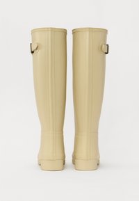 Botas de goma de color beige hasta la rodilla con una textura suave, correas traseras y costuras verticales. Diseño de punta redonda y suela plana.