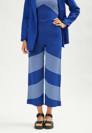 Pantaloni blu a maglia con gambe ampie presentano righe bianche orizzontali. Abbinati a un crop top blu e a un blazer blu. Sandali neri completano il look.