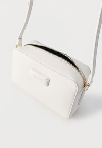 Άσπρη τσάντα crossbody Lacoste με χρυσό φερμουάρ και λογότυπο, με ρυθμιζόμενο λουρί και ορθογώνιο σχήμα σε λευκό φόντο.