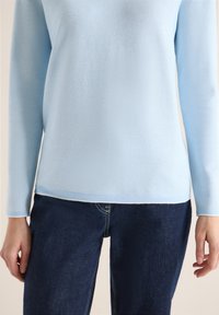 Personne portant un haut bleu clair texturé à manches longues et un jean en denim bleu foncé, mains détendues le long du corps sur un fond neutre.