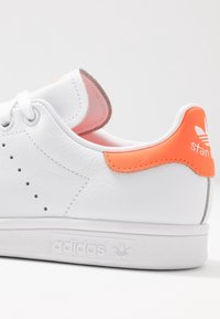White leather sneaker with orange heel tab, round toe, and perforated detailing. Features minimalistic design and textured sole with branding.