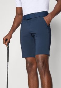 Námornícky modré golfové šortky vyrobené z flexibilného materiálu, s textúrovaným opaskom a vreckami. Navrhnuté pre pohodlie a pohyblivosť počas hry.