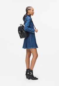Denim shirt dress with frayed hem; black leather backpack with a clasp; black ankle boots with silver hardware. Hair in braids and a scarf.