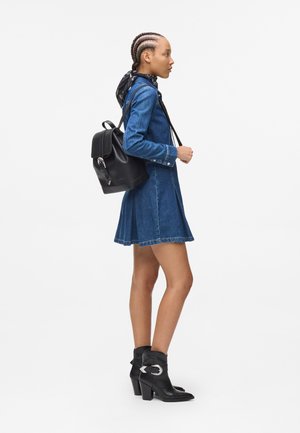 Denim shirt dress with frayed hem; black leather backpack with a clasp; black ankle boots with silver hardware. Hair in braids and a scarf.