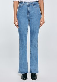 Osoba nosící světle modré džíny s vysokým pasem a rovnými nohavicemi, černé boty s špičatou špičkou a bílou košili zastrčenou dovnitř.