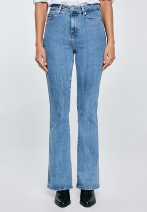 Inimene kannab helesiniseid kõrge vöökohaga sirgejalaseid teksaseid, musti terava ninaga saapaid ja valget sissepoole pistetud särki.