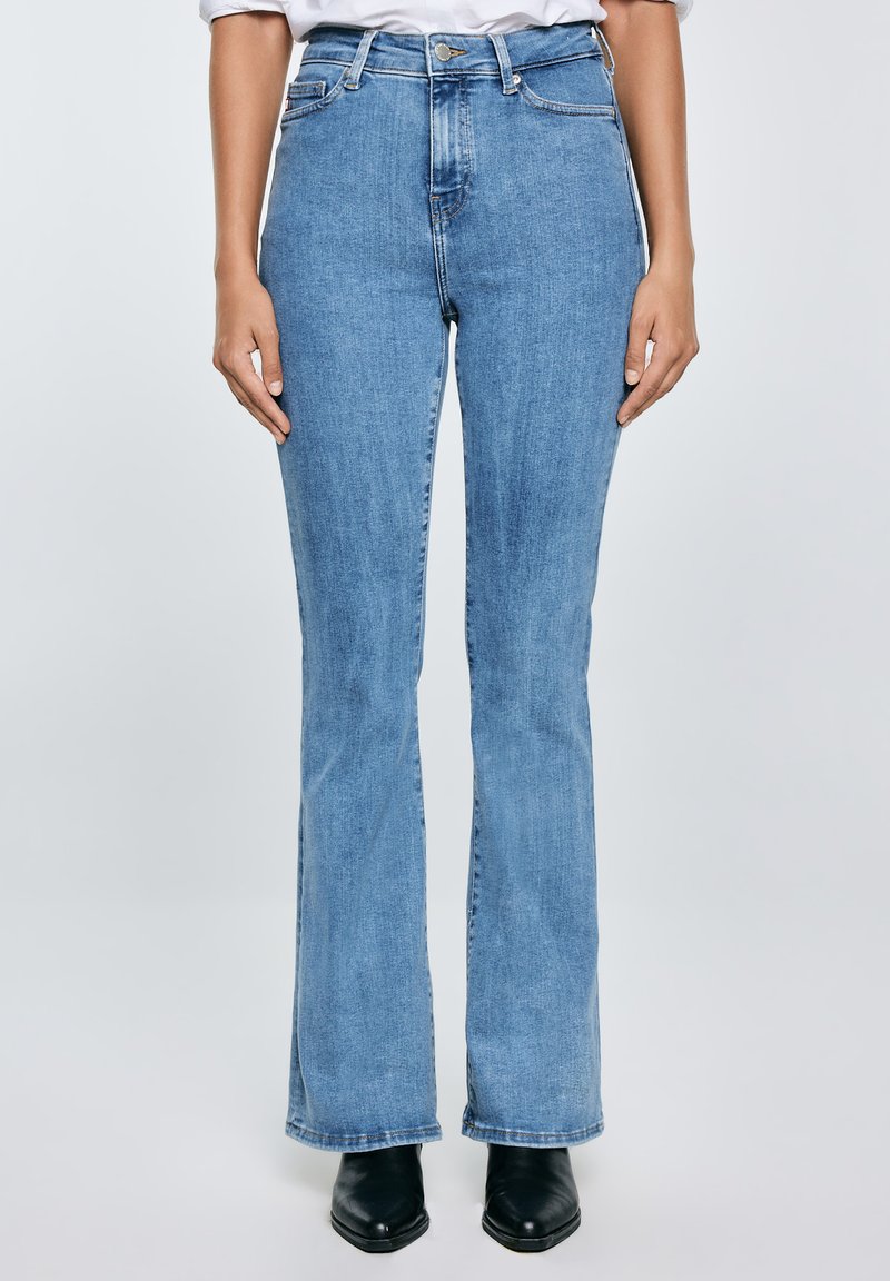 Osoba nosící světle modré džíny s vysokým pasem a rovnými nohavicemi, černé boty s špičatou špičkou a bílou košili zastrčenou dovnitř.