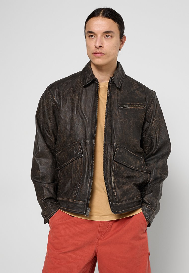 Brązowa skórzana kurtka o teksturowanym wykończeniu, z kieszonką na zamek i dwoma kieszeniami z przodu. Posiada kołnierz i widoczne szwy.