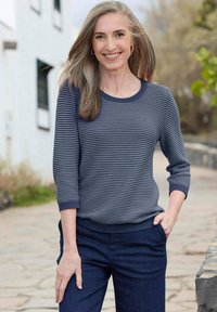Marine gestreifter Strickpullover mit Dreiviertelärmel und geripptem Ausschnitt, kombiniert mit dunkelblauen Hosen. Außensetting mit verschwommenem Hintergrund.